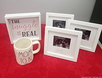 Photo showing a white framed sign with text 'The Snuggle Is Real', a white mug with black text 'Mom To Be', and three white square picture frames each holding an ultrasound photo placeholder labeled 'Hello Grandparents'.