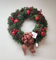 Full view of the circular Christmas wreath showing green artificial pine needles, brown pinecones, bright red berries, red apple ornaments, red shiny balls, and a plaid bow at the base.