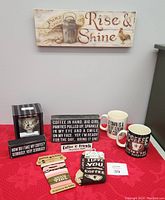 Photo of lot items on red tablecloth against gray wall: wall signs, coffee mugs, three coozies, teapot nightlight box.