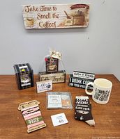 Photo shows assorted new coffee-themed items: wooden signs, coffee cup coozies, a white coffee cup, nightlight in packaging, and coffee-themed coasters arranged on table under a coffee sign on wall.