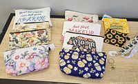 Photo showing 10 colorful cosmetic bags and purses with various inspirational, floral, and themed designs, along with a white lanyard and silver key ring laid on a table.