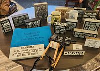 Photo of multiple new family gift giving items arranged on a table, including wooden signs, paddles, mugs, and a blue tote bag mostly themed around grandparents, moms, and dads.