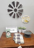 Photo of farmhouse-themed decor items on a brown wooden table against a gray wall, including a windmill-style clock, metal tray, kitchen scale, tea towels, faux plants, pedestal stand and a round sign.