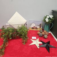 Photo of all farmhouse decor items on a red tablecloth, includes metal basket with greenery, embossed napkins, farm truck canvas, decorative metal stars, checkered spheres, and turquoise pitcher with flowers.