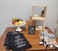 Photo shows full lot on a wooden table against grey wall. Includes faux fruit in white pedestal bowl, two wooden box shelves with chicken wire backing, wooden cutting boards, black tea towels with kitchen sayings, white decorative bowls, chalkboard-style sign, and spatulas still in packaging.