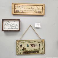 Photo showing three laundry room decorative signs hung on a grey wall with a small lot number card.