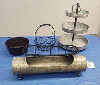 Five metal containers and racks displayed on a blue surface for decorating purposes: galvanized three-tier round tray, small round black metal bucket, oval wire basket with handle, long narrow wire basket, and tubular galvanized container with feet.