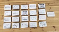 Top-down view of 25 mini rectangular wooden decorative signs arranged on a wooden floor. Each sign has unique inspirational or humorous quotes in various fonts, mostly black text on white background.