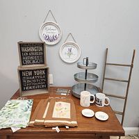 New wooden ladder, galvanized metal tiered tray, Rae Dunn mugs, decorative wooden signs, tea towels, small ceramic dishes with text, and hanging wall decor plates with lavender theme displayed on wooden table against light wall.