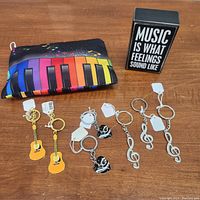 Photo showing a music-themed colorful zippered pouch with piano keys and musical notes, a black and white rectangular sign with a music quote, and six different music-inspired keychains: two yellow guitars with gold treble clefs, two silver treble clefs, and two black and white heart-shaped keychains.