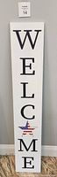 Full view of vertical white wooden welcome sign with large black letters and American flag star decoration.