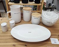 Full set of white stoneware items with black text and wooden lids on canisters, laid out on wooden table.