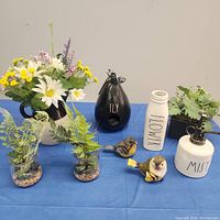 Photo shows Rae Dunn ceramic pieces: white vase labeled FLOWER, white mister labeled MIST, and black birdhouse labeled FLY, with two resin birds and multiple faux flower arrangements including daisies and greenery in white ceramic and glass jars with pebbles.