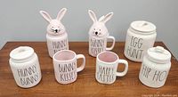 Photo of 7 ceramic Rae Dunn Easter themed items: 4 canisters with lids including two bunny head lids and three mugs with pink interiors, all labeled with Easter and bunny-themed text.