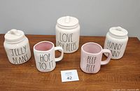 Five Rae Dunn ceramic items arranged on a wooden surface, consisting of three canisters and two mugs with Easter-themed text.
