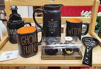 Photo showing 3 black ceramic Halloween mugs (one with haunted house lid) with orange interiors, a black pitcher with recipe text, a black spoon rest with white text, and boxed Halloween salt and pepper shakers.