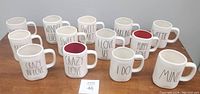 Photo of 13 new Rae Dunn ceramic mugs arranged on a table, each with a love-themed phrase in large black text on white matte ceramic. Includes some mugs with red interior.