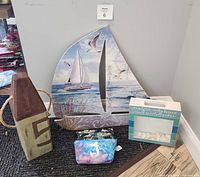 Photo showing four seashore themed items: large wooden sailboat wall decor, wooden buoy decor, decorative box with seashells, and ocean-themed cosmetic bag.