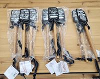 Nine new Primitives by Kathy spatulas with wooden handles and black silicone heads with white humorous text, individually wrapped in plastic and tied with black ribbons.