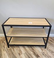 Three-tier wood and metal store display shelf with natural wood shelves and black metal frame, photographed against plain wall on wood-patterned floor.