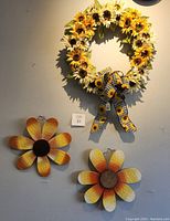 Photo showing a handmade sunflower wreath with a bold sunflower-print bow hanging on a wall, with two wooden sunflower-shaped decorations below it, all primarily yellow, orange, and brown.