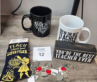 Photo shows two ceramic mugs with teacher-themed messages, three metal teacher key chains each with multiple charms including apples and hearts, a pair of black socks with yellow text, and a wooden block sign with teacher appreciation text.