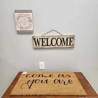Photo shows a doormat with 'come as you are' script in black on natural coir fiber, a wooden rectangular 'WELCOME' wall sign with black letters on white distressed background hanging above it, and a small floral decorative wall plaque with message 'Sweet Home' to the left.