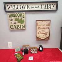Three cabin-themed wooden signs mounted on wall and table with black bear statues and road trip socks arranged on red cloth.