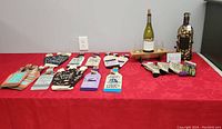Wide angle photo showing all items laid out on a red tablecloth, including multiple fabric wine bottle covers, cork holder, wooden stand with bottle and glasses, and wine socks.