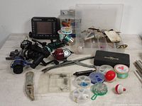 Lot of fishing items arranged on a table showing reels, Eagle fish finder, tackle boxes, fishing line, lures, and metal fish grippers.