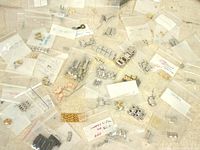 Top-down view showing multiple small plastic bags containing metal watch bracelet links and pins scattered on a surface. Bags are labeled with brand names and some model identifiers. Varied metal finishes include silver-tone, gold-tone, and rose gold-tone. Some bracelet segments have grid or link patterns.