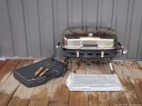 Portable stainless steel propane camping BBQ grill setup with folding legs, thermometer on lid, and a closed lid. Utensil case and bubble wrapped parts next to the grill.