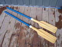 Two assembled paddles/oars with blue shafts and light brown blades laid on wooden floor showing full length and color.