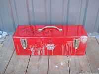 Front view of vintage red metal toolbox with paint drips and wear, closed with metal latches and central handle.