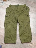 Front view of olive green vintage Canadian Forces cold weather windproof pants with multiple buttoned pockets, drawstring waist and ankle cuffs, laid flat on floor.