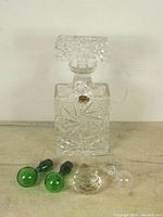 Clear pressed glass liquor decanter with faceted stopper and four additional glass bottle stoppers of varied design and colors on wooden surface.