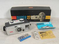 Full lot including camera, original box, sealed film roll, flashbulbs in packaging, flash guard, and paperwork laid out on surface.