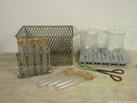 Full view of the vintage laboratory items arranged showing test tubes with cork stoppers, metal test tube rack, glass beakers in plastic rack, wire mesh basket, and metal tongs.
