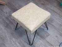 Overall angled view showing the neutral cream vinyl seat with thin brown atomic line pattern and black hairpin legs on a wood floor.