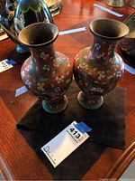 Two Asian inspired metal vases with floral enamel paint on a red base placed on a black cloth on a wooden table.