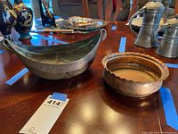 Photo showing two vintage metal bowls on a wooden table, one brass with script and an elongated oval shape, the other copper with scalloped rim.