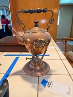 Front view of the vintage copper brass tea pot on its ornate stand and burner, showing the handle, lid, and round base.