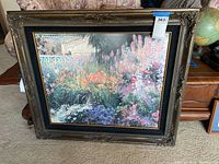Framed garden theme wall art poster standing on carpeted floor against furniture, showing full view of floral garden scene and decorative frame.