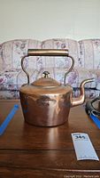 Large copper teapot with hinged lid, wooden handle grip, several dents, sitting on wooden table in front of couch.