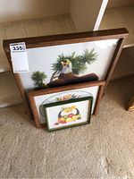 Framed needlepoint artworks and empty frame stacked leaning against wall, showing the scale and framing details