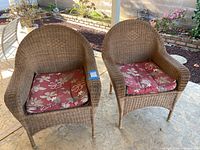Pair of two outdoor wicker patio chairs with floral red cushions placed on stone flooring outdoors.