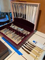 Wooden case holding various silver plated flatware pieces including knives with cream handles, forks, spoons, and serving utensils all arranged in maroon velvet slots