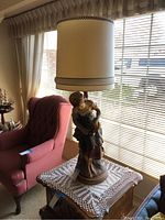 Full view of the vintage statue lamp with beige drum shade, positioned on a side table next to a maroon armchair.