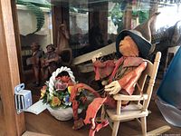 Wood carved figurines displayed in a glass cabinet including a horse head sculpture, two men with musical instruments, fabric scarecrow in a rocking chair, and ceramic flower basket.