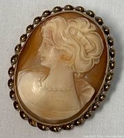 Front view of oval cameo brooch pendant showing carved female bust with detailed hair and necklace against brown background, surrounded by twisted rope gold filled frame.